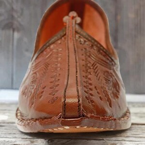 May include: A brown leather slip-on shoe featuring an upturned pointed toe and intricate embossed floral patterns across the upper surface. The shoe has a central stitched seam running down the front and a visible thick sole with edge stitching.