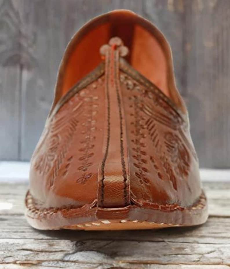 May include: A brown leather slip-on shoe featuring an upturned pointed toe and intricate embossed floral patterns across the upper surface. The shoe has a central stitched seam running down the front and a visible thick sole with edge stitching.