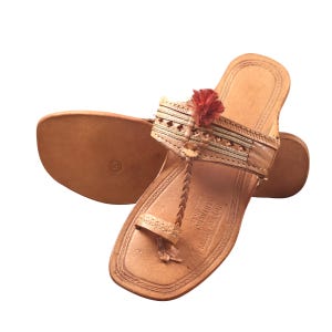 May include: Tan leather sandals with a braided toe strap and decorative accents. The sandals feature a small red pom-pom and intricate cut-out designs on the strap. The sole is a light brown color.