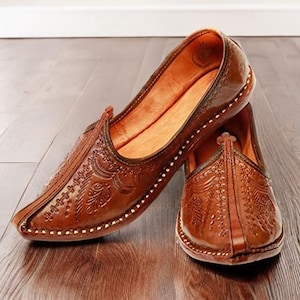 May include: A pair of brown leather slip-on shoes featuring intricate embossed floral patterns across the upper surface. The shoes have a pointed toe design, a low profile, and visible stitching along the edges of the sole and the central seam.