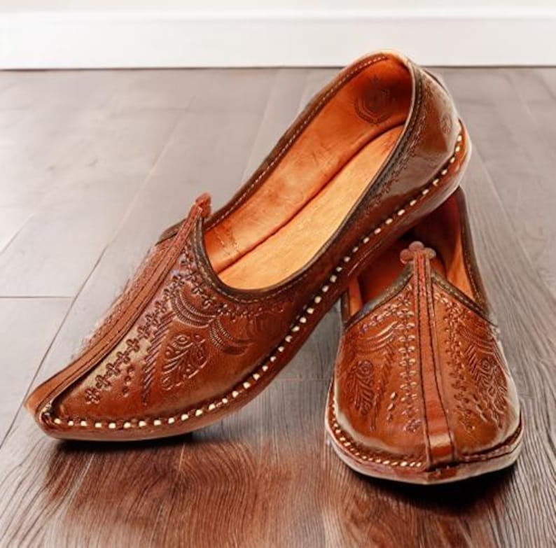 May include: A pair of brown leather slip-on shoes featuring intricate embossed floral patterns across the upper surface. The shoes have a pointed toe design, a low profile, and visible stitching along the edges of the sole and the central seam.