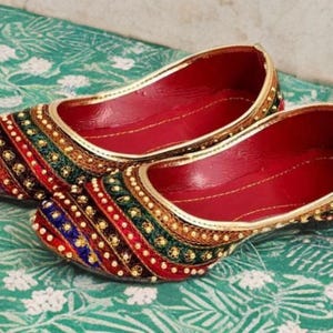 May include: A pair of slip-on flats featuring a vibrant multi-colored upper with red, green, blue, and gold embroidered stripes accented by small white beads. The interior of the shoes is lined in a solid red material with gold trim edges.