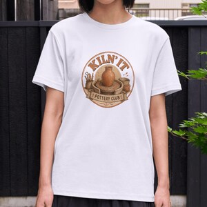 Puede incluir: Una camiseta blanca de manga corta con un gráfico circular en el pecho. El gráfico muestra un jarrón de arcilla marrón sobre un torno de alfarero con el texto KILN' IT POTTERY CLUB en letras marrones, rodeado de pequeñas herramientas.