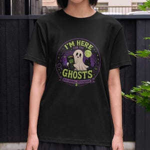 May include: Black t-shirt with a circular graphic featuring a cartoon ghost holding a device. The text reads "I'M HERE FOR THE GHOSTS" and "PARANORMAL INVESTIGATOR". The design includes a full moon and trees.