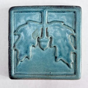 May include: A square ceramic tile featuring a relief design of two oak leaves and a central acorn. The tile has a glossy, variegated blue-green glaze with darker edges and a smooth, raised border framing the botanical motif in the centre.