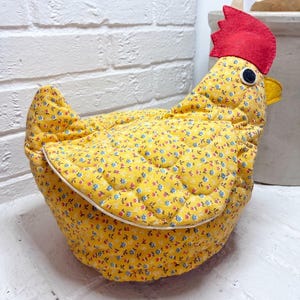 May include: A quilted fabric chicken ornament featuring a bright yellow base with a small blue and red floral pattern. The chicken has a prominent red felt comb on its head, a yellow felt beak, and a circular black and white eye detail.