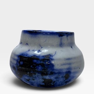 May include: A small, rounded ceramic vase featuring a white base glaze with deep blue and black mottled, dripped patterns. The vessel has a wide, bulbous body that tapers toward a short, straight neck with a dark blue rim and a smooth finish.