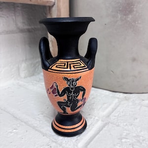 Puede incluir: Un pequeño jarrón de cerámica negro y naranja con una ilustración de estilo de figuras negras de una criatura similar a un sátiro. El jarrón tiene dos asas, un borde acampanado y una base con un patrón geométrico griego en el cuello.