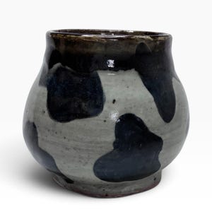 May include: A small, rounded ceramic vessel featuring a glossy, light gray base glaze adorned with irregular, dark navy blue patches. The rim is finished with a dark, solid navy blue band, and the overall surface has a smooth, reflective texture.