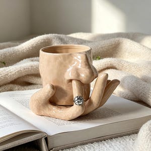 Könnte beinhalten: Ein beigefarbener Keramikbecher mit einer modellierten menschlichen Nase auf der Vorderseite und einem Sockel in Handform, dessen Finger zur Nase reichen. Ein silberfarbener Ring mit texturierter, unregelmäßiger Oberfläche steckt am Zeigefinger.