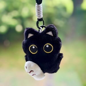 Op de afbeelding: Een zwarte pluche kattenhanger met grote, geel omrande ogen en een witte poot onder de kin. De kat heeft kleine puntige oortjes met witte details aan de binnenkant en is bevestigd aan een zwart koord met een wit gewikkeld accent en een clip.