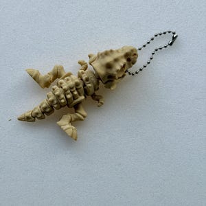 Puede incluir: Un llavero con forma de esqueleto de dinosaurio de color beige que presenta un cráneo detallado, una caja torácica articulada y una cola larga. La figura está fabricada en plástico moldeado y unida a una cadena de bolas de metal plateado.