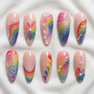 Pode incluir: Uma coleção de dez unhas artificiais em forma de amêndoa, cada uma adornada com designs exclusivos com tema de arco-íris. As unhas apresentam uma variedade de enfeites, incluindo pérolas e detalhes florais, com uma paleta de cores rosa, azul, amarelo e verde.