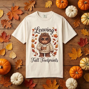 May include: A cream-colored short-sleeve t-shirt featuring a graphic of a brown Bigfoot wearing a beige cable-knit sweater, leopard print headband, and sunglasses while holding a coffee cup, with the text Leaving Fall Footprints.