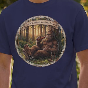 May include: A navy blue t-shirt featuring a circular graphic of a brown Bigfoot sitting in a forest, drinking from a bottle. A banner above the creature displays the text, "They still haven't found me...losers." The design has a weathered, vintage look.