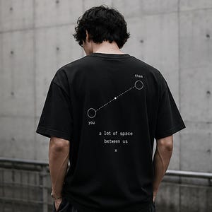 May include: A black short-sleeved t-shirt featuring a white graphic on the back. The design displays two circles connected by a dashed line, labelled 'you' and 'them', with the text 'a lot of space between us' and an 'x' printed below.