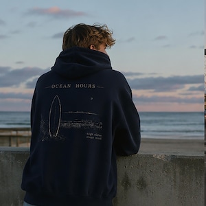 May include: A navy blue hooded sweatshirt featuring a white graphic on the back. The design includes a surfboard, a small crescent moon, and the text "OCEAN HOURS" above the graphic with "high tides clear mind" printed below the surfboard.