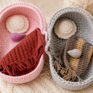May include: Two oval crochet baskets, one light pink and one light gray, each containing a round cream-colored crochet pillow. The pink basket holds a rust-colored fringed blanket and a small purple crochet teardrop. The gray basket holds a brown and tan striped fringed blanket and a small beige crochet teardrop.
