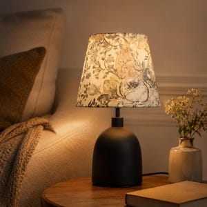 May include: A black table lamp with a floral patterned lampshade. The lampshade features a cream background with pink, green, and gray floral designs. The lamp sits on a round wooden table next to a vase with flowers and a closed book.