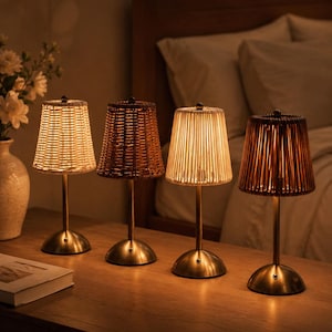 May include: Four small table lamps with brass bases and different shade designs. The shades are made of woven rattan, ribbed glass, and woven brown material. The lamps are on a wooden surface, with a bed and flowers in the background.