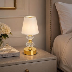 May include: A table lamp featuring a white tapered fabric shade, a gold-toned metal base, and two stacked clear faceted crystal spheres. The lamp emits a soft glow, highlighting the reflective gold finish and the intricate cut of the crystal.