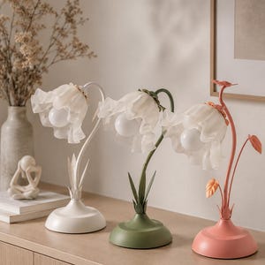 Puede incluir: Tres lámparas de mesa con forma de flor en blanco, verde y coral. Cada lámpara tiene una pantalla translúcida y con volantes y una bombilla. Las bases son redondas y están coloreadas para que coincidan con los tallos. Las lámparas son decorativas.