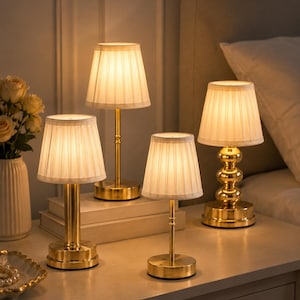 May include: Four gold-toned table lamps with pleated white shades are arranged on a nightstand. The lamps have varying base designs, including a stacked ball and a simple round base. A vase of flowers and a decorative dish are also visible.