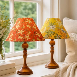 May include: Two wooden table lamps with floral patterned shades. One shade is orange with white and yellow flowers, the other is yellow with white and green flowers. The lamps sit on a white windowsill.