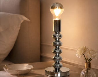 May include: A chrome table lamp with a large light bulb. The lamp has a stacked, spherical base and sits on a round base. A small white bowl, a book, and a vase with white flowers are on the table.