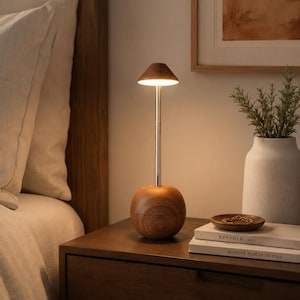 Könnte beinhalten: Eine tragbare Tischleuchte mit einem kugelförmigen Holzsockel mit sichtbarer Maserung, einem schlanken, silberfarbenen Metallschaft und einem konischen Holzschirm, der durch eine LED ein warmes, nach unten gerichtetes Licht abgibt.