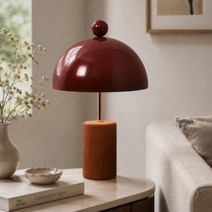May include: A modern table lamp with a burgundy dome shade and a wooden base. The lamp features a brass stem and a small burgundy sphere on top. The lamp is on a marble-topped table, with a book and small dish nearby. The lamp is a decorative home decor item.