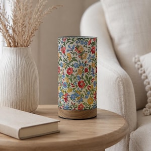 May include: A cylindrical lamp with a floral pattern in red, blue, and yellow on a cream background. The lamp sits on a wooden base and is placed on a round wooden side table. A vase with dried plants and a book are also on the table.