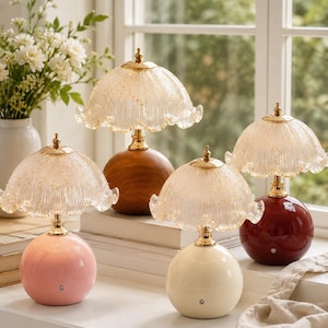 May include: A collection of decorative table lamps with a vintage aesthetic. Each lamp features a round base in various colors, including pink, cream, and burgundy, topped with a gold-toned stem and a glass shade with a scalloped edge.