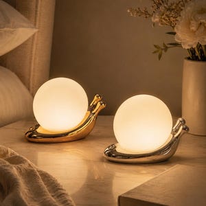 May include: Two decorative table lamps shaped like snails, each with a glowing spherical light. One lamp is gold, the other silver, both resting on a marble surface. A book and a folded blanket are also visible.