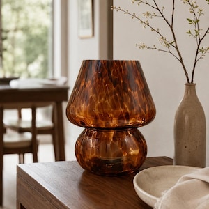 May include: A decorative amber glass vase with a unique mushroom-like shape. The vase sits on a wooden table, alongside a ceramic bowl and a small vase holding a flowering branch. The glass has a speckled pattern.