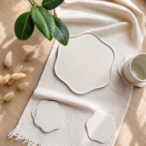 Könnte beinhalten: Ein cremefarbenes Keramik-Geschirrset, bestehend aus einem großen Teller, einer Tasse und mehreren Untersetzern, angeordnet auf einem strukturierten, cremefarbenen Stoff. Das Set hat eine organische Form. Grüne Blätter und getrocknete Pflanzen sorgen für einen natürlichen Touch.