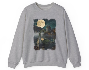 May include: A heather grey crewneck sweatshirt featuring a graphic print of a long-necked creature emerging from dark water at night. The scene includes a full moon, a stone castle on the shore, and misty mountains in the background.