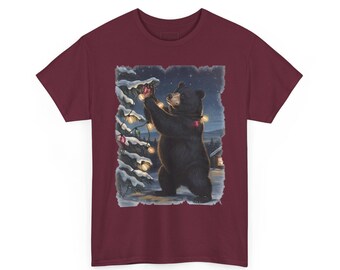 May include: A maroon short-sleeve t-shirt featuring a graphic print of a black bear standing on its hind legs, decorating a snow-covered pine tree with glowing string lights in a winter forest setting at night.