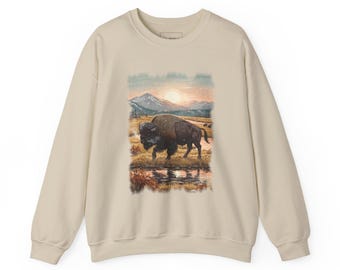 May include: A beige crewneck sweatshirt featuring a graphic print of a brown bison standing in a grassy field near a stream. The background depicts a mountain range under a bright sun, with soft colours and a painterly style throughout the scene.