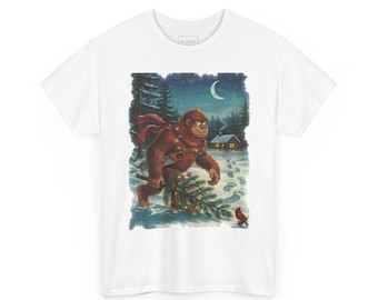 May include: A white t-shirt featuring a graphic illustration of a brown Bigfoot creature wearing a red scarf and wrapped in string lights, carrying a decorated pine tree through a snowy forest at night with a cabin and crescent moon.