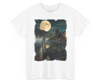 May include: A white short-sleeved t-shirt featuring a graphic print of a long-necked green creature swimming in a dark lake at night. The scene includes a full moon, a stone castle on the shore, and a small lit cabin under a dark sky.