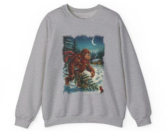 May include: A heather grey crewneck sweatshirt featuring a graphic print of a brown Bigfoot creature wearing a red scarf and carrying a decorated Christmas tree through a snowy forest at night under a crescent moon near a small cabin.