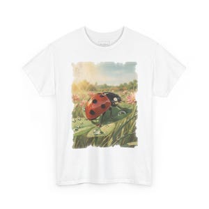 Può includere: Una t-shirt bianca a maniche corte con una stampa grafica rettangolare centrale raffigurante una coccinella rossa con punti neri su una foglia verde coperta di gocce d'acqua, su uno sfondo sfocato di erba e fiori rosa.