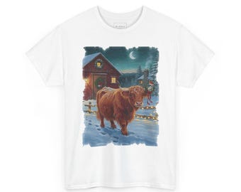May include: A white t-shirt featuring a graphic illustration of a brown Highland cow standing in deep snow in front of a red barn at night. The scene includes a crescent moon, a lit fence, and a wreath on the barn door, creating a winter setting.