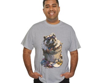 May include: A heather grey short-sleeved t-shirt featuring a graphic illustration of a raccoon wearing dark sunglasses while sitting inside a rubbish bin. The raccoon is surrounded by various pieces of colourful debris and discarded items at the base.