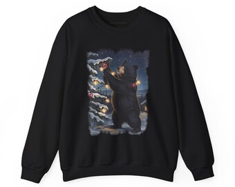 May include: A black crewneck sweatshirt featuring a graphic print of a black bear standing on its hind legs, decorating a snow-covered pine tree with glowing string lights in a dark, starry winter forest setting at night.