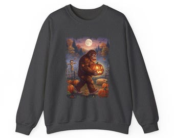 May include: A dark grey crewneck sweatshirt featuring a graphic print of a brown, furry creature walking through a pumpkin patch at night. The scene includes a glowing jack-o'-lantern, a scarecrow, a full moon, and autumn trees in the background.