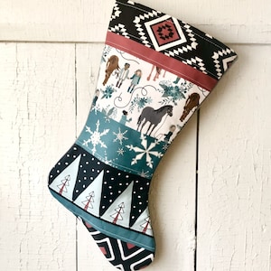 May include: A Christmas stocking with a patchwork design featuring a winter scene with people and horses, snowflakes, and trees. The stocking is made of fabric with a black, white, blue, and red color scheme.