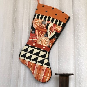May include: A Christmas stocking with a festive design, featuring orange, black, and white patterns. The stocking is decorated with ornaments and geometric shapes. Also, an antique iron, wooden spools, and a tall wooden spool holder are on display.
