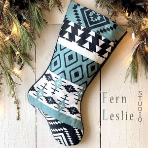 May include: A Christmas stocking with a teal, black, and white geometric pattern. The stocking is hanging against a white wooden background, with evergreen branches and lights in the background. The text "Fern Leslie Studio" is visible.